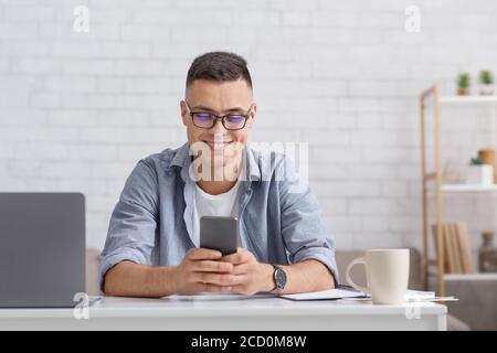 Tippen Sie auf Ihrem Telefon und arbeiten Sie online. Lächelnder Kerl im Hemd schaut auf Smartphone, sitzt am Tisch mit Laptop Stockfoto