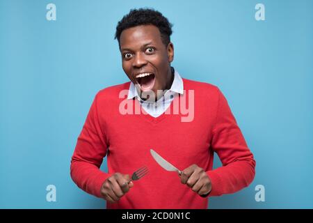 Hungriger afrikanischer Mann, der Gabel und Messer hält und auf das Abendessen wartet. Stockfoto