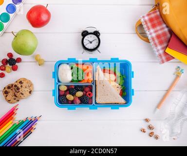 Offene Lunchbox mit Sandwich, Gemüse, Ei, frischen Beeren auf dem weißen Holzhintergrund in der Nähe der Uhr, Schulzubehör und Rucksack. Draufsicht, FLA Stockfoto