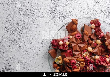 Gebrochene Tafeln Milchschokolade mit Nüssen und sublimierten Beeren und Schokolade Bonbons in der Platte auf texturiertem grauen Hintergrund, mit leerem Platz für te Stockfoto