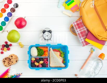 Offene Lunchbox mit Sandwich, Gemüse, Ei, frischen Beeren auf dem weißen Holzhintergrund in der Nähe von Uhr und Schulzubehör. Draufsicht, flach liegend. Stockfoto