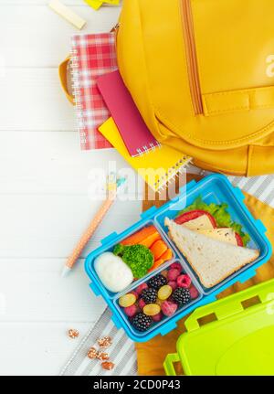 Offene Lunchbox mit Sandwich, Gemüse, Ei, frischen Beeren auf dem weißen Holzhintergrund in der Nähe von Schulzubehör und Rucksack. Draufsicht, flach liegend. Stockfoto