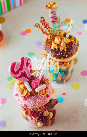 Draufsicht auf zwei Freak-Shakes mit Donut, Popcorn und Süßigkeiten auf Party-Hintergrund; selektiver Fokus auf Himbeer-Freak-Shake. Stockfoto