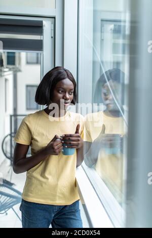 Elegante afroamerikanische Frau, die nachdenklich aus dem großen Fenster schaut Während sie ihre Tasse Tee in den Händen hielt Stockfoto