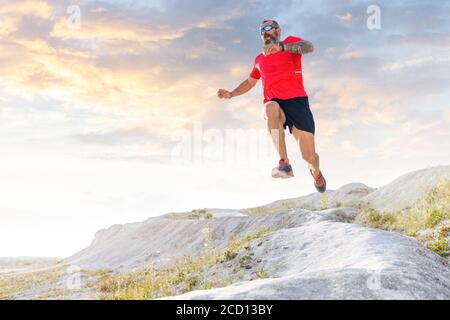 Bärtiger Läufer springt von der Schanze auf Trailrunning Cross. Der reife Sportler läuft Ultra Trail in der Wüste gegen den malerischen Abendhimmel Stockfoto