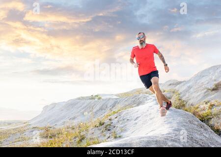 Bärtiger Läufer springt von der Schanze auf Trailrunning Cross. Der reife Sportler läuft Ultra Trail in der Wüste gegen den malerischen Abendhimmel Stockfoto