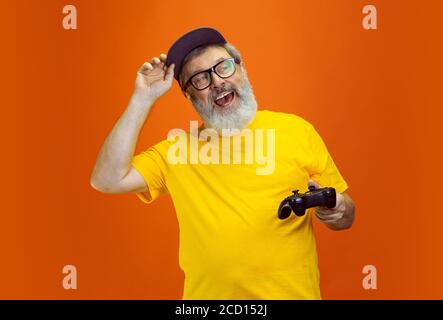 Gamer Mann. Portrait von Senior Hipster Mann mit Geräten, Gadgets isoliert auf orangen Studio-Hintergrund. Tech und fröhliche ältere Lifestyle-Konzept. Trendige Farben, für immer Jugend. Copyspace für Ihre Anzeige. Stockfoto
