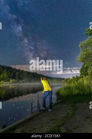 Ein Mann zeigt auf die Milchstraße des Nachthimmels und lehnt sich an ein Geländer vor einem See. Stockfoto