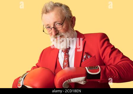 Senior Business man trägt roten formalen Anzug und roten Boxhandschuhe Blick auf Kamera, während in der Verteidigung Pose auf gelbem Hintergrund stehen. Wirtschaftliche Komp Stockfoto