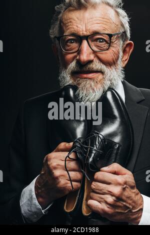 Glücklich elegante ältere Mann in Anzug Blick auf die Kamera mit Freude und Aufregung hält fest neue Lederschuhe in der Nähe seiner Brust, schätzt seine teuer Stockfoto