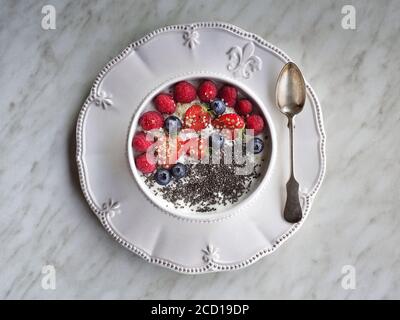 Joghurt mit Beeren (Blaubeeren, Erdbeeren und Himbeeren) und Chiasamen in weißer Schüssel auf weißem Teller und weißem Hintergrund. Overhead-Aufnahme. Stockfoto