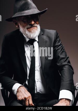 Elegant gekleidete ältere Gentleman Portrait im klassischen Stil in Trilby Hut und schwarzem Anzug. Männermode, klassisches Look Konzept. Studio im Dunkeln Stockfoto