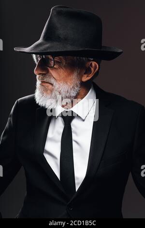 Elegant gekleidete ältere Gentleman Portrait im klassischen Stil in Trilby Hut und schwarzem Anzug. Männermode, klassisches Look Konzept. Studio im Dunkeln Stockfoto