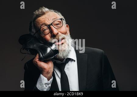 Studio Portrait von positiven graubärtigen alten Mann in schwarzem Anzug, weißes Hemd und schwarze Krawatte halten neben Kopf maßgeschneiderte schwarze Schuhe, als ob es ein Griff o Stockfoto