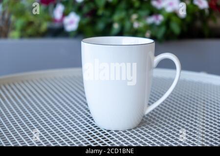 Kaffeetasse Mockup auf einem Gartentisch. Weiße Farbe leere Tasse mit Griff für heiße Getränke, Blur blühenden Blumen Hintergrund. Werbung, Branding-Vorlage Stockfoto