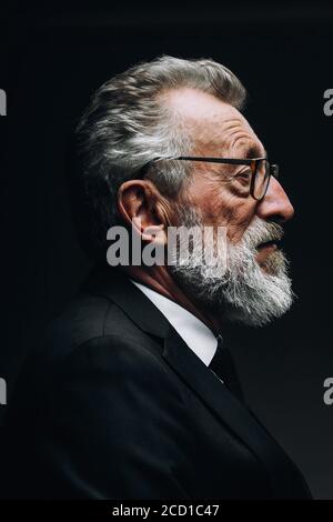 Profil-Headshot des intelligenten bärtigen grauhaarigen Wissenschaftler hören Bericht, gekleidet in formalen schwarzen Anzug, sitzen isoliert über dunkel Stockfoto