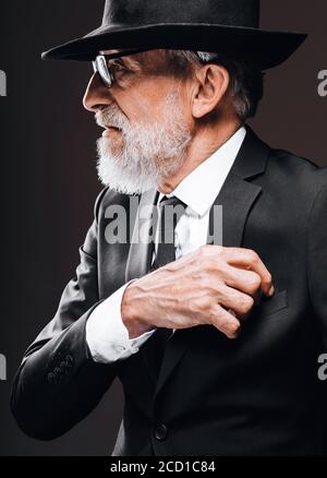 Altgealterter bärtiger Mann im Bild eines englischen Geheimagenten, der schwarzen Anzug mit Hut auf den Augen trägt und vor dunklem Hintergrund posiert. Stockfoto