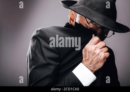 Ein alter männlicher Schauspieler, der eine englische Spionagerolle spielt, in schwarzem Anzug gekleidet, versteckt hinter Fedora-Hut, mit geheimnisvollem eleganten Look. Isoliertes Studio portra Stockfoto
