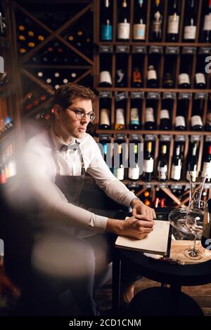 Verschwommene Aufnahme eines nachdenklichen Sommeliers, der in einem Notizblock einige Dinge schrieb, die zwischen einer sehr großen Auswahl an Weinen in den Regalen des Weinrestaurants saßen. Stockfoto