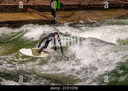 River-Surfing auf dem Eisbach in München, Deutschland. Die stehende Welle kann so lange gesurft werden, wie das Gleichgewicht hält, und in geschäftigen Zeiten bildet sich eine Schlange von Surfern am Ufer Stockfoto