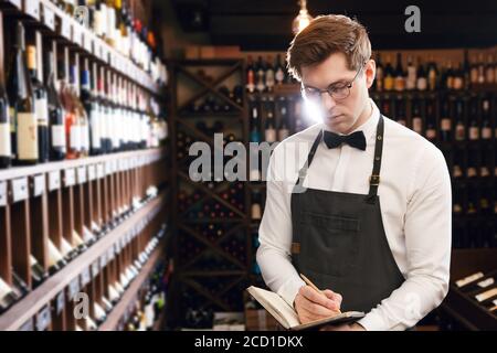 Professioneller männlicher Kavist, der sich Notizen bei der Degustationskarte in der Nähe gemacht hat Weinregale der europäischen Wein boutiuque Stockfoto