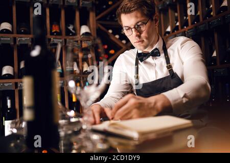 Nachdenklicher männlicher Sommelier mit Blick auf eine sehr breite Palette von Weinen aus den wichtigsten Regionen der Welt, die von Weinregalen aufgestellt wurden, und in Notizblock einige Th Stockfoto