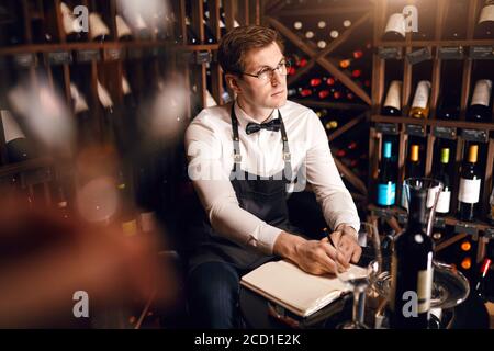 Verschwommene Aufnahme eines nachdenklichen Sommeliers, der in einem Notizblock einige Dinge schrieb, die zwischen einer sehr großen Auswahl an Weinen in den Regalen des Weinrestaurants saßen. Stockfoto