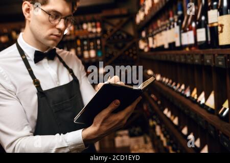 Professioneller männlicher Kavist, der sich Notizen bei der Degustationskarte in der Nähe gemacht hat Weinregale der europäischen Wein boutiuque Stockfoto