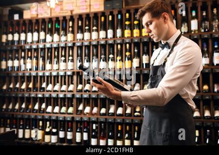 Professioneller Weinverkäufer oder Höhlenforscher, der eine Flasche Rotwein anbietet, Mann in weißem Hemd mit Fliege und Schürze, der über die Herkunft dieses oder erzählt Stockfoto