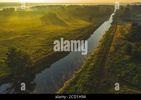 Luftaufnahme über ein Tal mit Nebel bei Sonnenaufgang. Drohne fliegt über Sommerfluss, Wiese und Wald. Die Strahlen der Sonne machen ihren Weg durch den Nebel Stockfoto