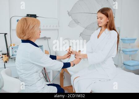 Junge hübsche kaukasische Frau, die auf dem Bett liegt und ihre sympathische Krankenschwester im Krankenhauszimmer isoliert, um den Gesundheitszustand zu untersuchen Stockfoto