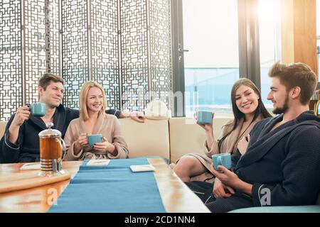 Entspannt diverse Freunde in Bademänteln entspannen in der Lounge Zone der Badehaus, trinken heißen Tee und die gemeinsame Nutzung positiver Eindrücke Stockfoto