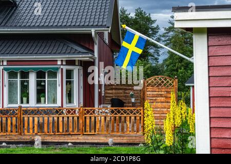 Vorderansicht des traditionellen schwedischen hölzernen roten Hauses mit Sommerterrasse auf dem Land. Schwedische Flagge auf der rechten Seite, montiert auf kleines Gästehaus, summe Stockfoto