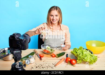 Fröhliche junge blonde Köchin präsentiert kulinarische Talente, nimmt an TV-Kochshow, trägt bequemes Hemd und Schürze, posiert am Kit Stockfoto