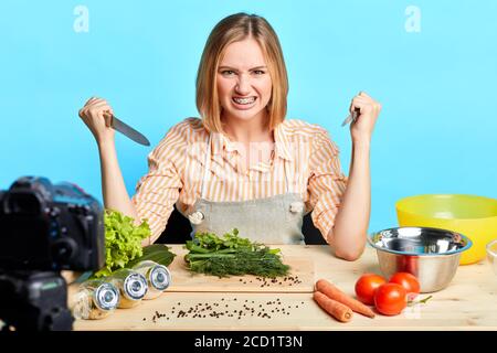 Blonde weibliche Köchin hält zwei Messer in den Händen, hat wütend und verrückt Gesichtsausdruck, so als ob sie angreifen, spielen Narr und Scherz mit Sho Stockfoto