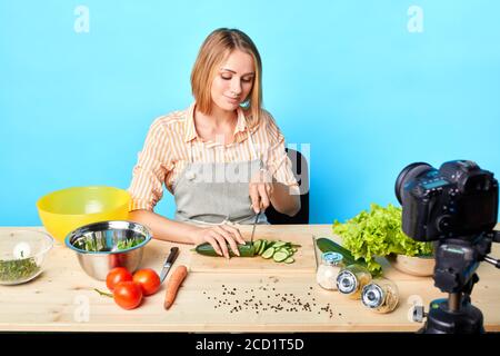 Studio-Aufnahme von schönen jungen Bloggerin schneiden frische rohe Gurke für Salat, Aufnahme neuer Inhalte für ihre vegetarische Lebensmittel Blog, genießt Kochen PR Stockfoto