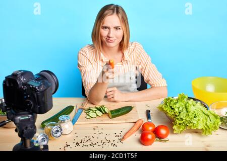 Frontansicht der verspielten hübschen weiblichen Köchin Punkte mit roher Karotte an der Kamera, hat freudige und schlaue Gesichtsausdruck, Kochen frische vegetarische Gerichte, angezogen Stockfoto