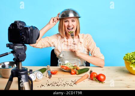 Erfolgreiche glückliche Köchin scherzen und spielen während Online-Kochkurs Master, posiert in Casual Shirt mit Streifen, Schürze und mit Schüssel auf ihr er Stockfoto