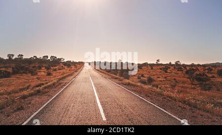 Legendäre RN7 - steht für Route Nationale - Straße durch wilde afrikanische Savanne mit kleinen Bäumen und Büschen an den Seiten, Sonne scheint im Hintergrund, t Stockfoto