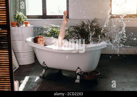 Entspannende Frau mit Spaß hebt ihre schlanken Beine in der Luft, während Schaumbad zu Hause. Wasserspritzer und fröhliche Stimmung. Spa- und Entspannungskonzept. Stockfoto