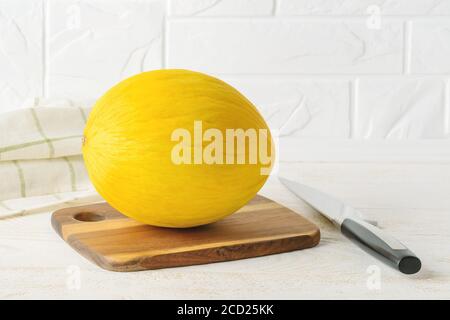 Ganze gelbe reife Honigmelone auf einem braunen Holzbrett und großes Messer über einem weißen Küchentisch. Vegetarisch, Rohkost, gesunde Ernährung. Stockfoto