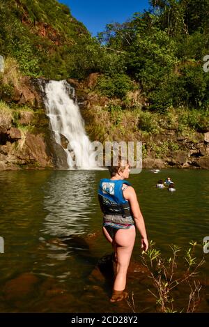 Frau im String Badeanzug, trägt Rettungsschwimmer, watend ins Wasser, um unter 45-Fuß-Wasserfall zu schwimmen, Waimea Valley, Oahu Island, Haleiwa, Hawaii, USA Stockfoto