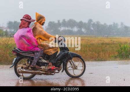 Kampot, Kambodscha - 14. Dezember 2016: Zwei Personen (nicht identifiziert) in rosa und orangen Plastikregenmänteln fahren bei starkem Regen ein Motorrad. Stockfoto
