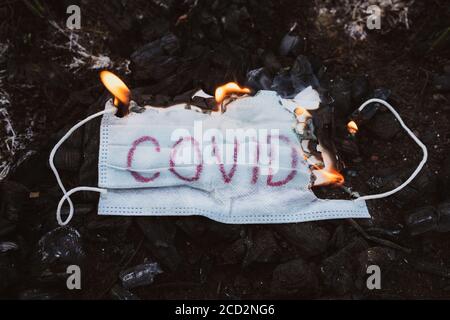 Medizinische Maske auf schwarzem Hintergrund mit der Aufschrift covid-19. Verbrennung Zerstörung der schützenden Gesichtsmaske. Verschmutzung der Natur durch covid m Stockfoto