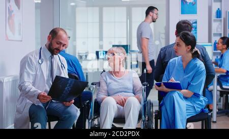 Arzt mit Stethoskop hält behinderte ältere Frau Radiographie im Rollstuhl, während mit ihr im Krankenhaus Wartebereich sprechen. Patient fragt nach seiner Ernennung an der Klinikrezeption. Stockfoto