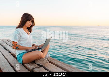 Eine junge schöne Frau mit Tattoos sitzt auf einem Pier am Meer und arbeitet an einem Laptop. Meer und Himmel im Hintergrund. Speicherplatz kopieren. Konzept der RE Stockfoto