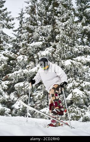 Skifahrer macht Tricks in den Bergen in der Wintersaison. Mann Freerider macht Sprung beim Rutschen nach schneebedeckten Pisten mit schönen schneebedeckten Fichtenwald im Hintergrund. Seitenansicht. Stockfoto