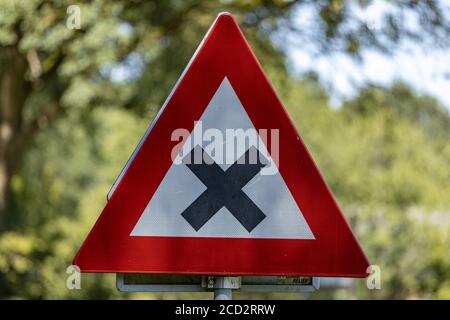 HOGE HEXEL, NIEDERLANDE - 14. Aug 2020: Holländisches Verkehrszeichen rotes Dreieck bedeutet eine gefährliche Überquerung mit unscharf Laub von Bäumen in Th Stockfoto