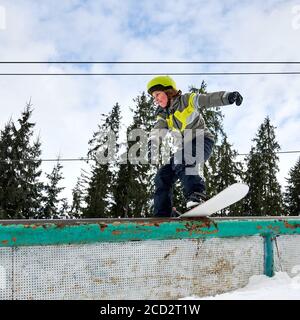 Kinder Snowboarder im Helm Reiten Snowboard unter schönen bewölkten Himmel mit Pinien im Hintergrund. Boy Performing Tricks mit Snowboard. Konzept des Snowboardsports und der Wintersport-Aktivitäten. Stockfoto