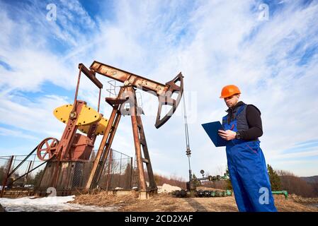 Petroleum Ingenieur in Arbeit Overalls und Helm hält Klemmbrett, Überprüfung Öl-Pumpeinheit, Notizen machen. Ölarbeiter steht in der Nähe von Ölpumpenheber unter schönem Himmel. Konzept der Ölförderung Stockfoto